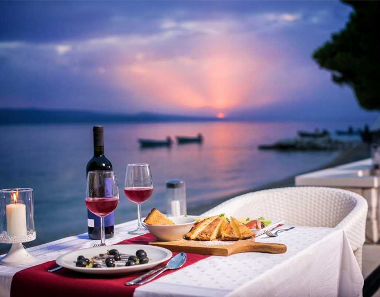 Table with appetizers, two glasses of wine, and candles at a romantic waterfront dinner at sunset, showcasing Miami Premier Transportation’s luxury evening services